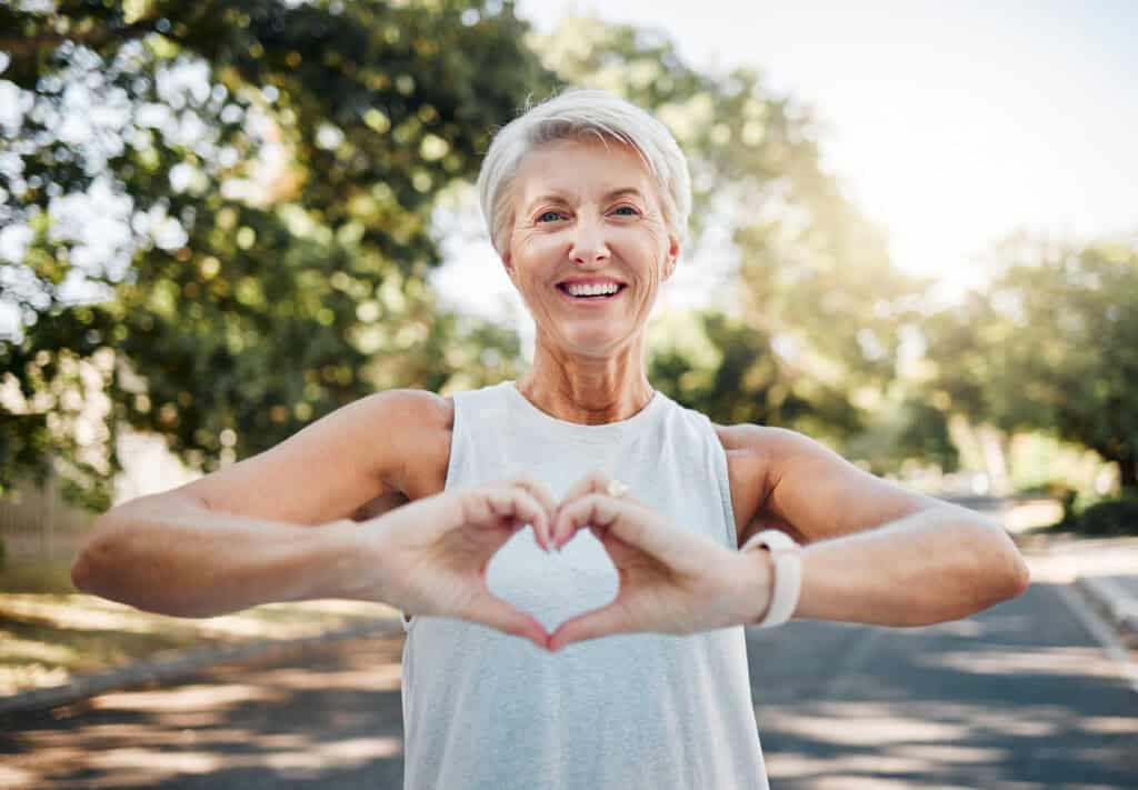 Smiling elderly woman outdoors, forming a heart shape with her hands toward the camera.