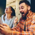 Two friends laugh while taking a selfie at a table, each holding a smartphone near a bright indoor setting diy style.
