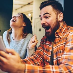 Two friends laugh while taking a selfie at a table, each holding a smartphone near a bright indoor setting diy style.