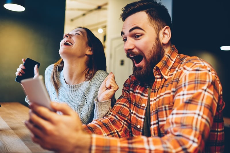 Two friends laugh while taking a selfie at a table, each holding a smartphone near a bright indoor setting diy style.