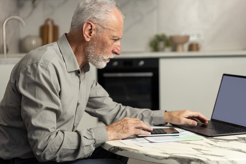 Older man with gray hair and beard sits at a kitchen table, using a calculator beside a laptop and financial papers.