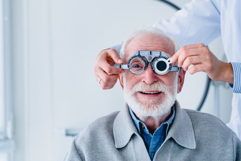 Older man smiles during an eye exam as a clinician adjusts a phoropter in front of his eyes.